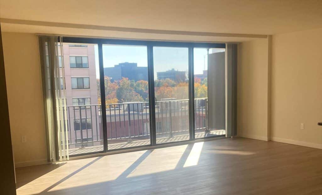 Empty room with light wood flooring, large floor-to-ceiling windows, a balcony, and a view of city buildings and trees in the background.