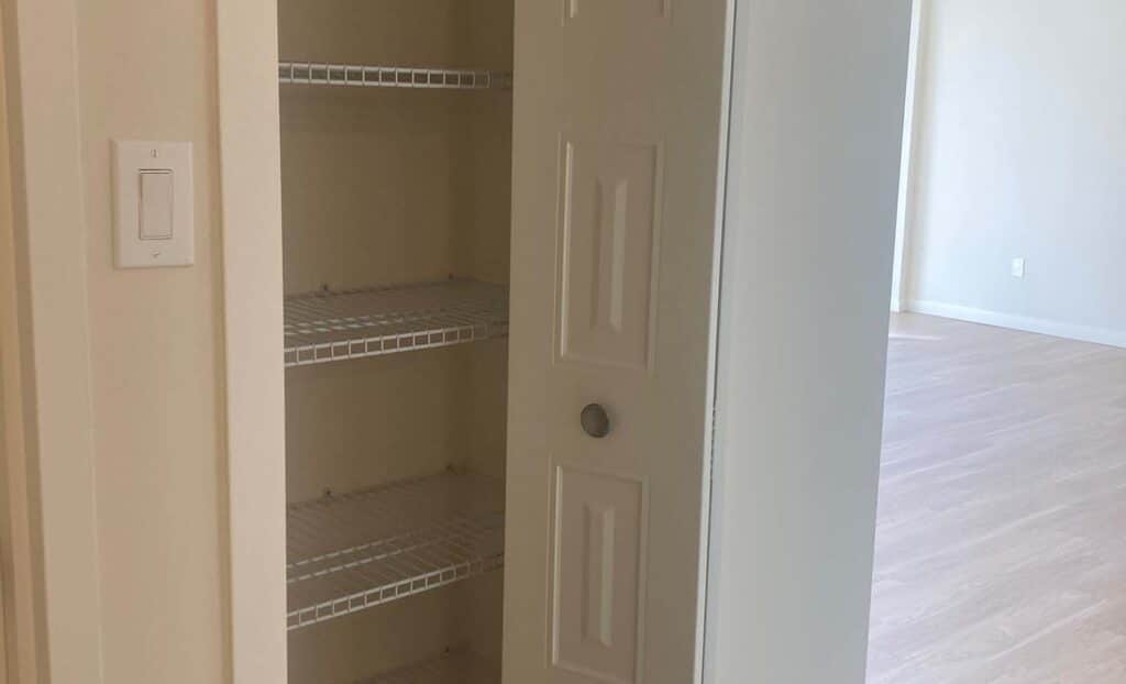 An open closet with four empty wire shelves and a white sliding door, located next to a light switch and a room with light wood flooring.