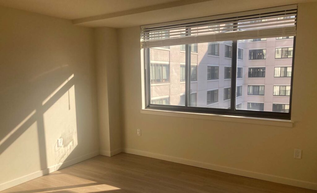 Empty room with light wood floor, beige walls, a large window with blinds, and sunlight casting shadows on the wall; city building visible outside.