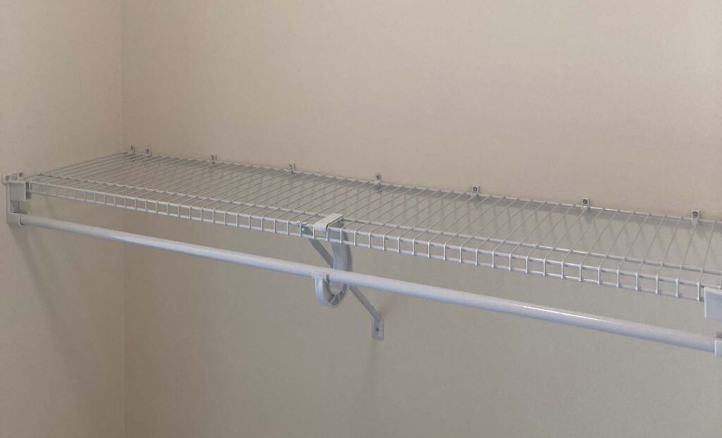 A white metal wire closet shelf with a built-in hanging rod is mounted on a beige wall.