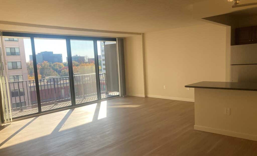 A sunlit, empty apartment with large floor-to-ceiling windows, a balcony, light wood flooring, and a kitchen with a stainless steel refrigerator.
