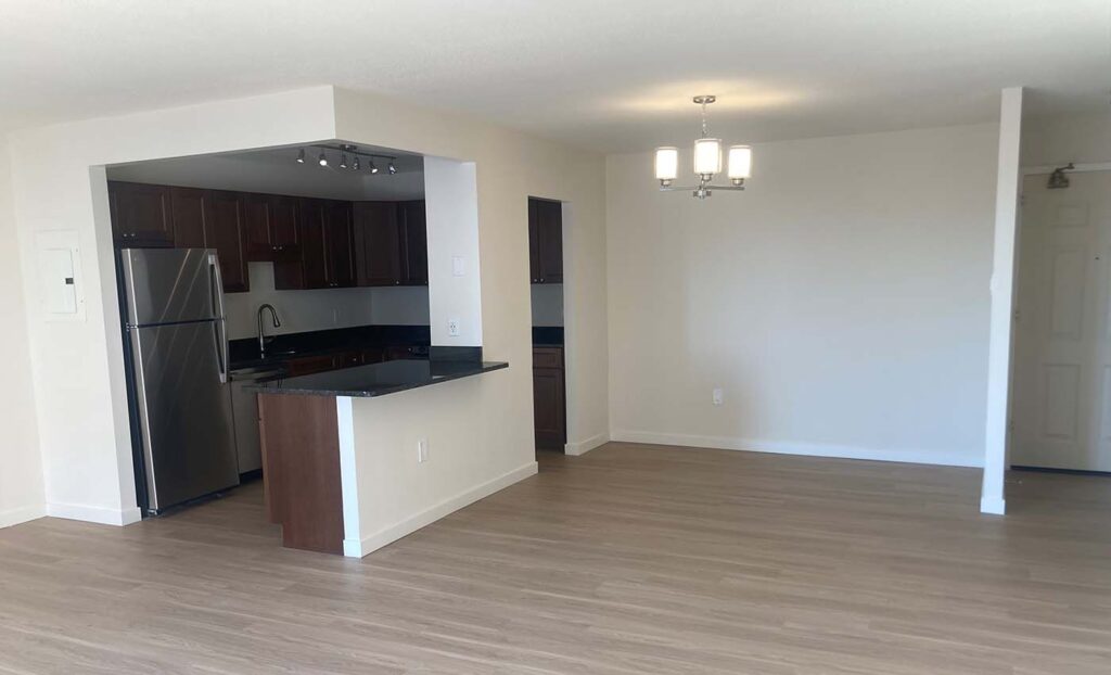 Unfurnished apartment interior with an open kitchen featuring stainless steel appliances, dark cabinets, and a breakfast bar; adjacent dining area with light fixture and wood flooring.