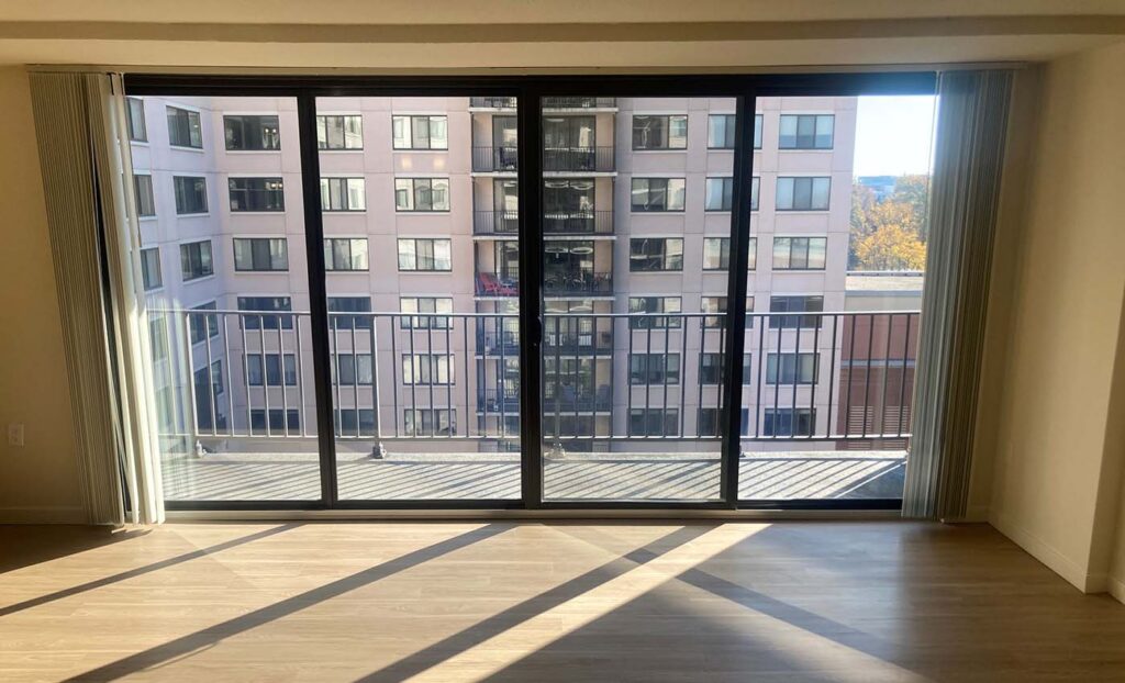 A large glass sliding door opens to a balcony with metal railing, overlooking a neighboring apartment building; sunlight casts shadows on the floor.