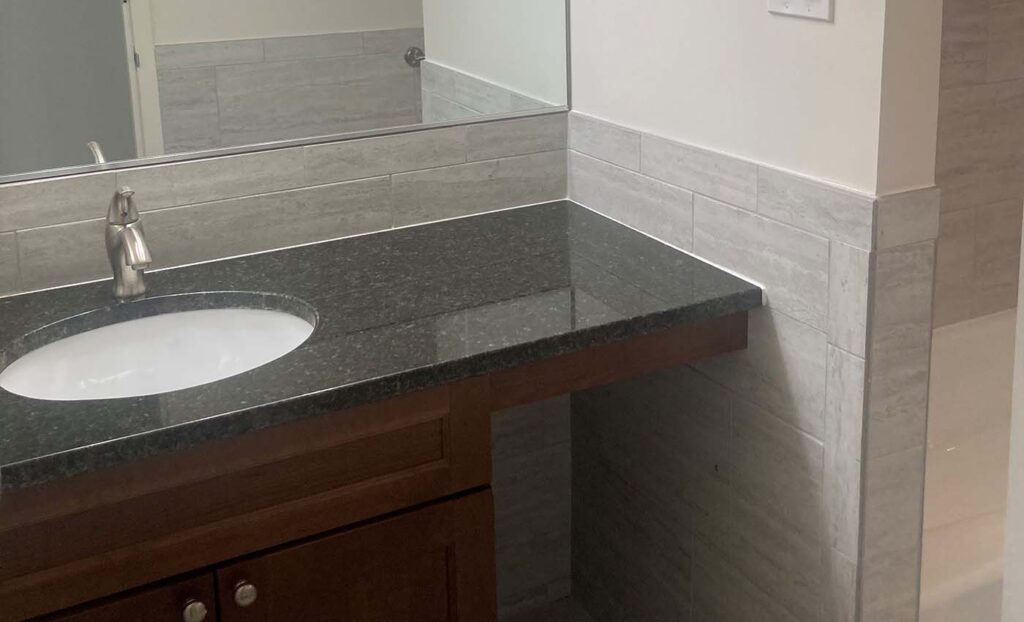Bathroom vanity with a dark countertop, built-in sink, wooden cabinet, large wall mirror, and light-colored tiled walls.