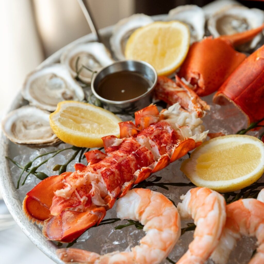 A seafood platter with lobster tail, shrimp, oysters, lemon wedges, and a small dish of sauce served on crushed ice.
