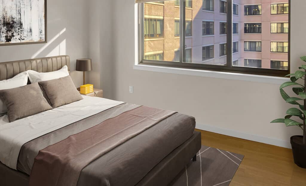 A neatly made bed with brown and beige bedding sits next to a window overlooking an apartment building; a plant and lamp are in the corner.