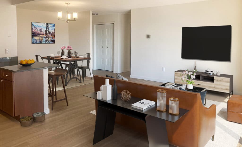 Open-plan living and dining area with modern furnishings, a wall-mounted TV, dining table, and kitchen island with fruit bowl.