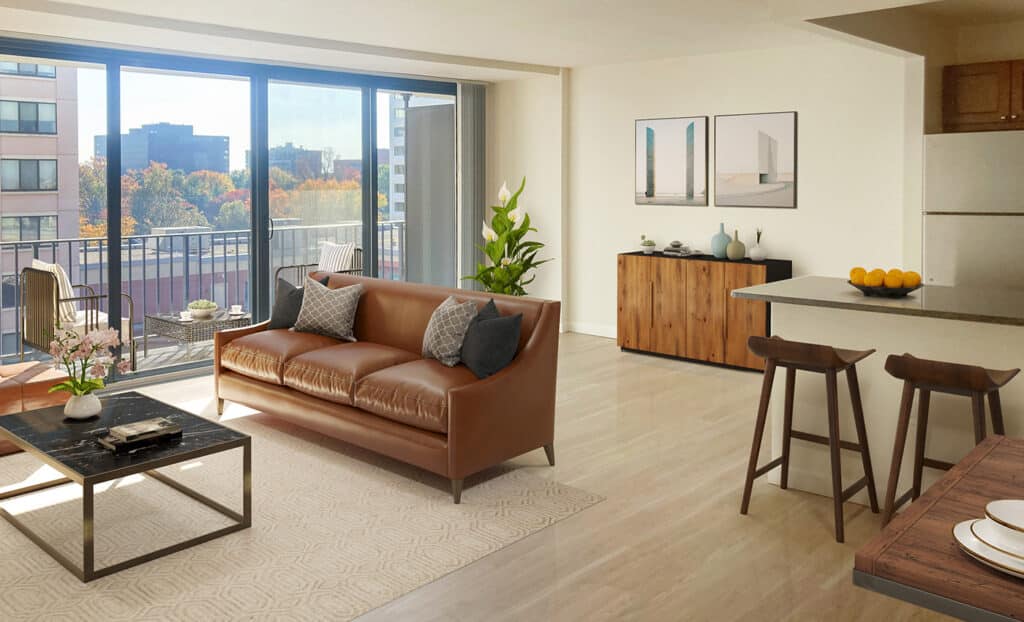 Modern apartment living room with brown leather sofa, coffee table, barstools at kitchen counter, wooden sideboard, large windows, and balcony view of buildings and trees.