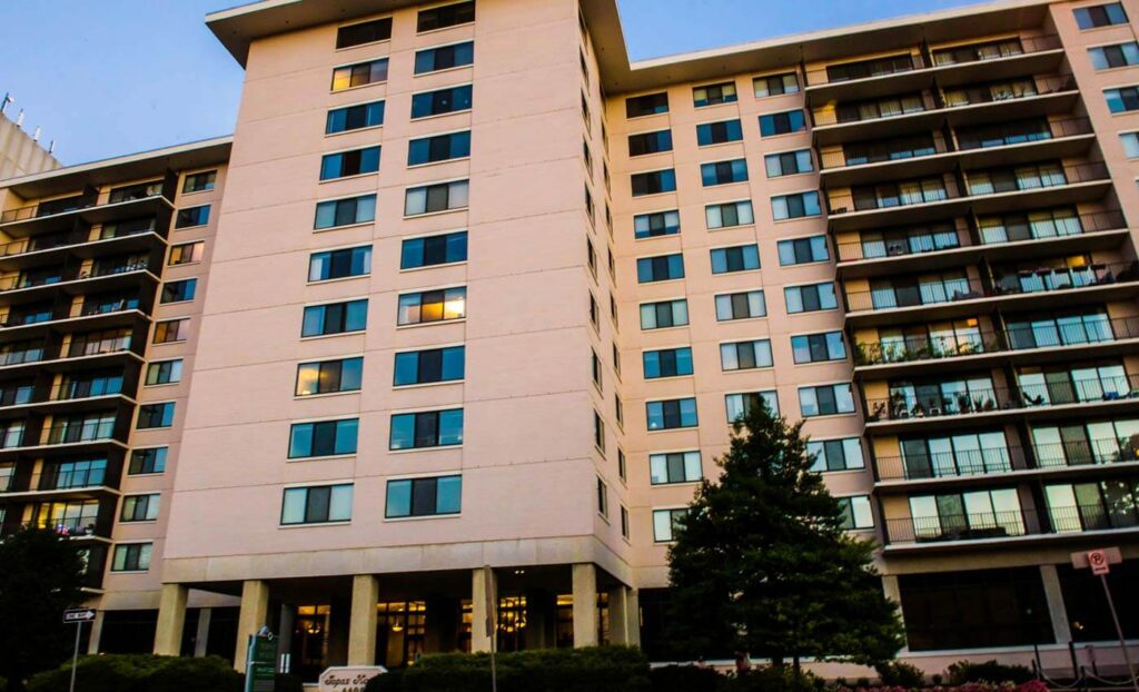 A multi-story apartment building with numerous windows and balconies, surrounded by trees and bushes, is shown in daylight.