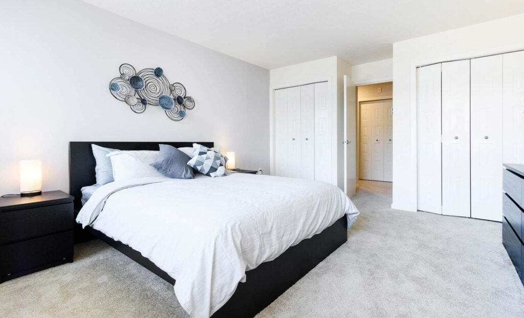 Modern bedroom with a neatly made bed, two nightstands with lamps, decorative wall art above the headboard, and white closets in the background.