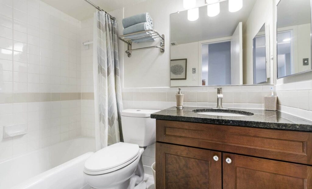 A clean bathroom with a bathtub and shower, a toilet, a dark wood vanity with a sink, mirror, and shelves holding towels above the toilet.