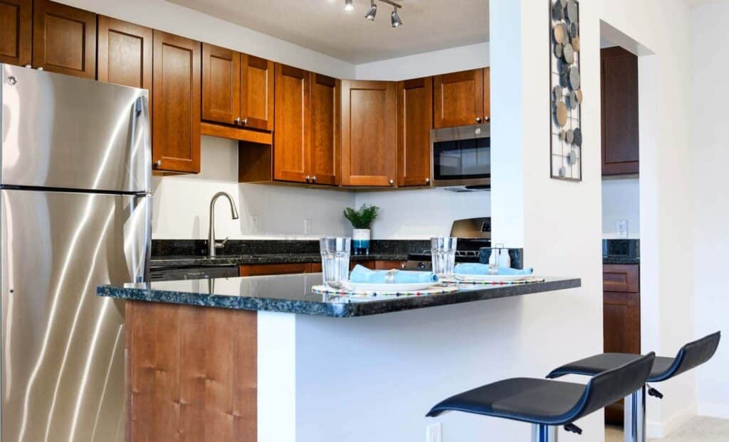 Modern kitchen with wooden cabinets, stainless steel appliances, black granite countertops, a small potted plant, and two black bar stools next to a breakfast bar.