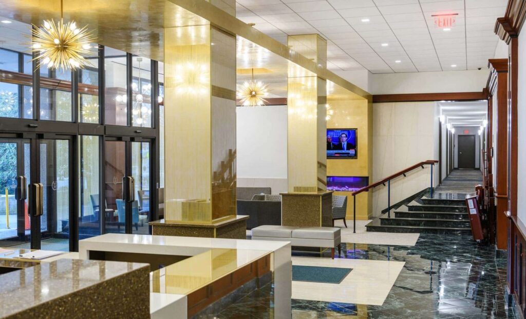 Modern hotel lobby with polished floors, gold columns, contemporary furniture, large windows, chandelier lighting, and a TV mounted on the wall near a hallway.