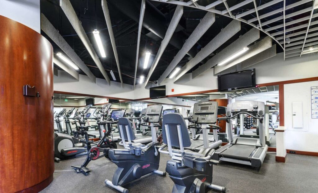 A gym with several treadmills, stationary bikes, and ellipticals in front of mirrors, under a ceiling with exposed beams.