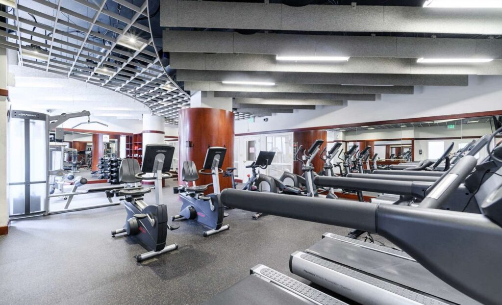 A modern gym with treadmills, stationary bikes, and various exercise machines, featuring large mirrors and ceiling panels.