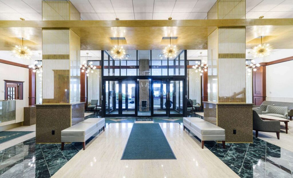 Spacious building lobby with glass entrance doors, gold ceiling accents, modern chandeliers, marble floors, and silver benches.