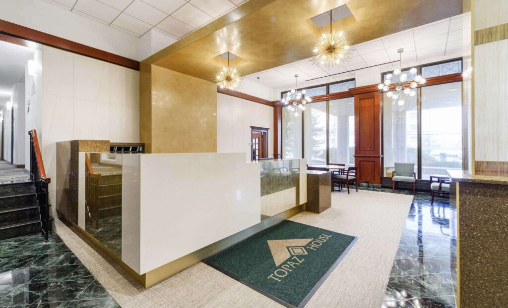 Modern office lobby with a white reception desk, gold ceiling accents, geometric light fixtures, and a green rug labeled "TOPAZ HOUSE." Several chairs are by large windows in the background.