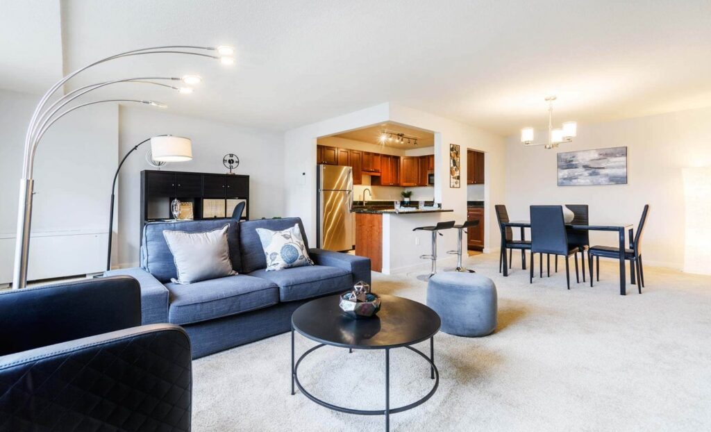 Modern open-concept living and dining area with a blue sofa, round coffee table, dining table for six, and kitchen with stainless steel appliances in the background.