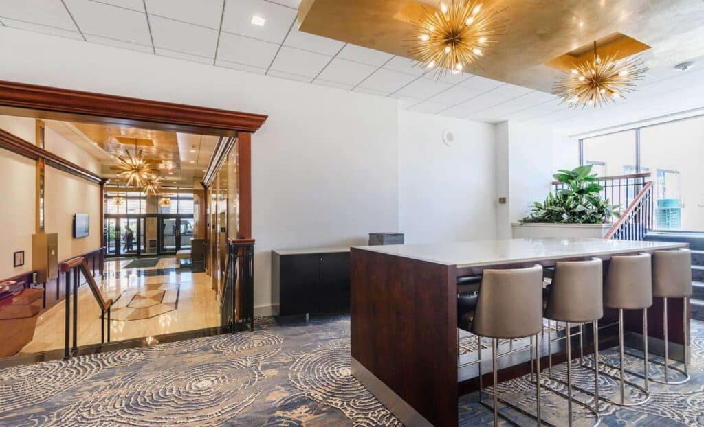 Modern lounge area with a high table and chairs, decorative lighting fixtures, patterned carpet, plants, and large windows letting in natural light.