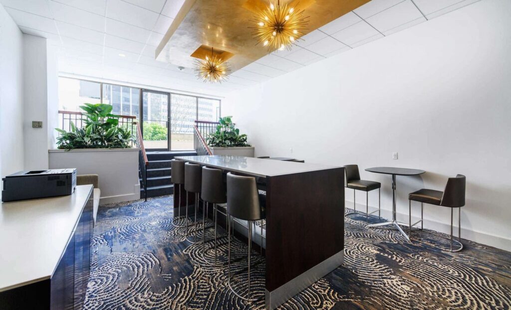 Modern office lounge with a high table, chairs, a small round table, plants, a printer, and large windows letting in natural light.