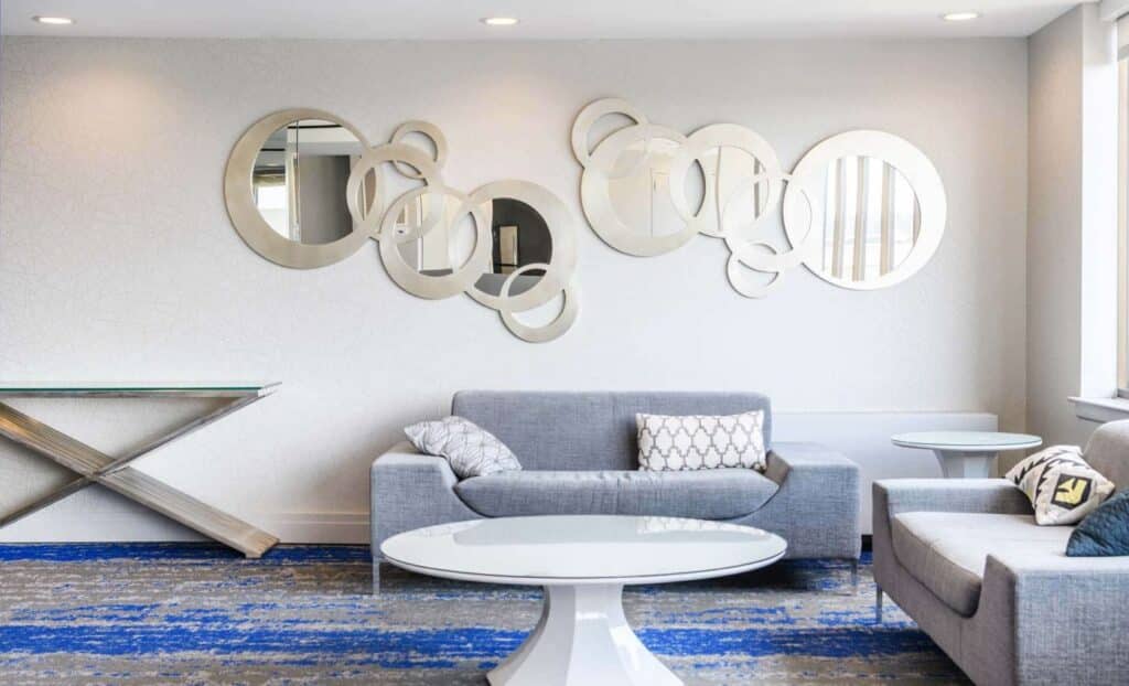 Modern lounge area with gray sofas, round white coffee tables, decorative pillows, and circular mirrors on a white wall. Blue and gray carpet covers the floor.