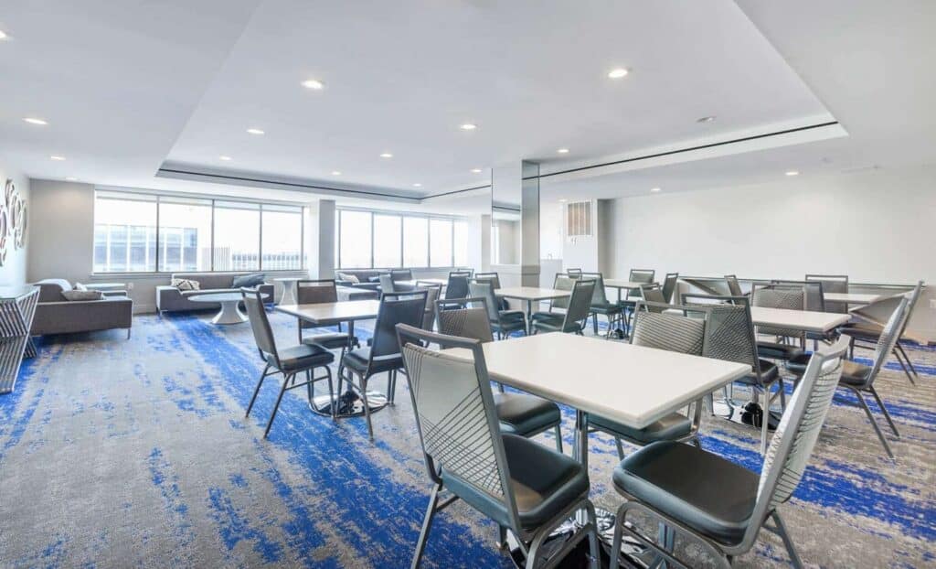 A modern, brightly lit room with several tables and chairs, blue-patterned carpet, large windows, and seating areas along the sides.