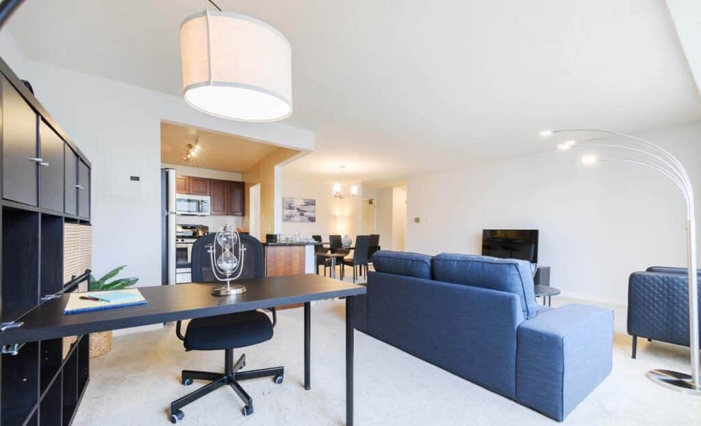 Bright, modern open-plan living space with a blue sofa, black desk and chair, dining area, and kitchen in the background.
