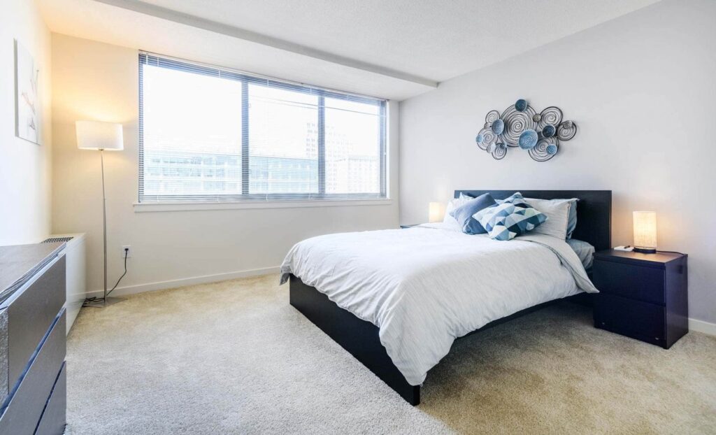 A bright bedroom with a large window, white walls, a double bed with white bedding, two nightstands with lamps, a dresser, and modern wall art above the bed.