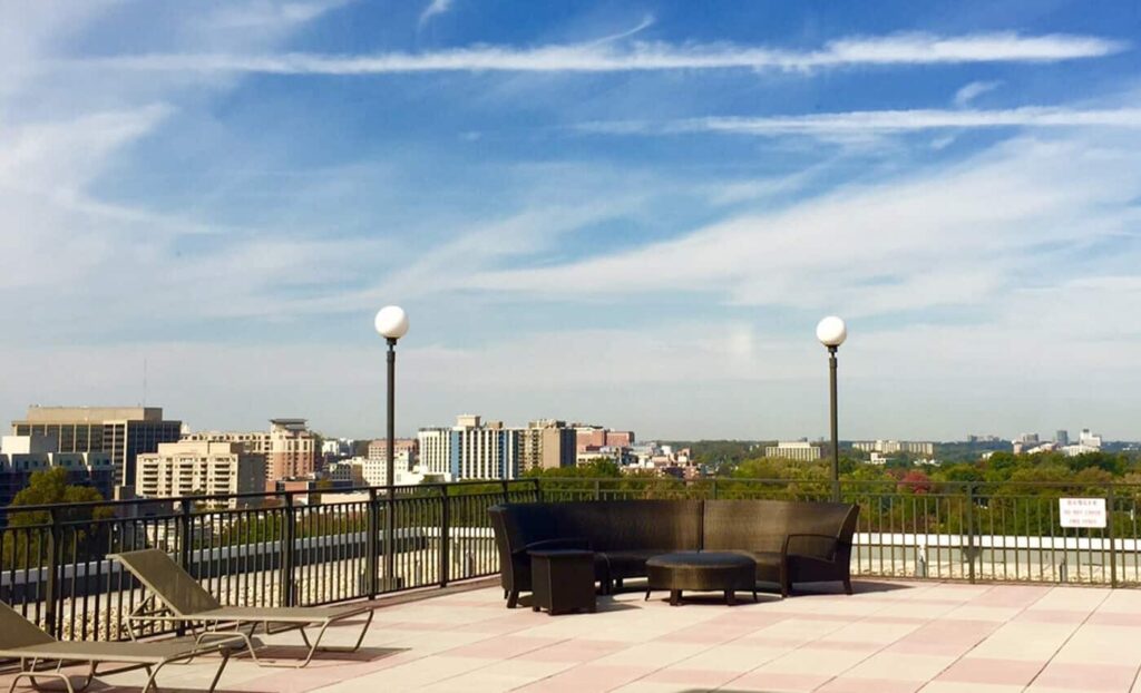 Rooftop patio with lounge chairs, outdoor seating, and city buildings in the background under a blue sky with clouds.