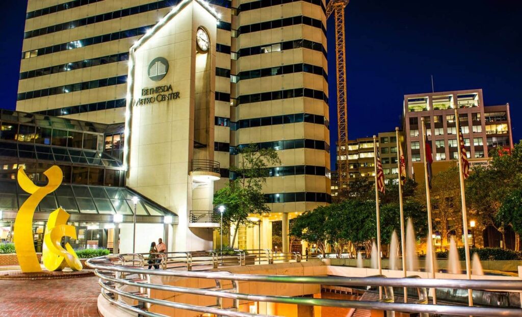 Night view of Bethesda Metro Center with illuminated office buildings, a clock tower, public art sculptures, and a fountain in the plaza.
