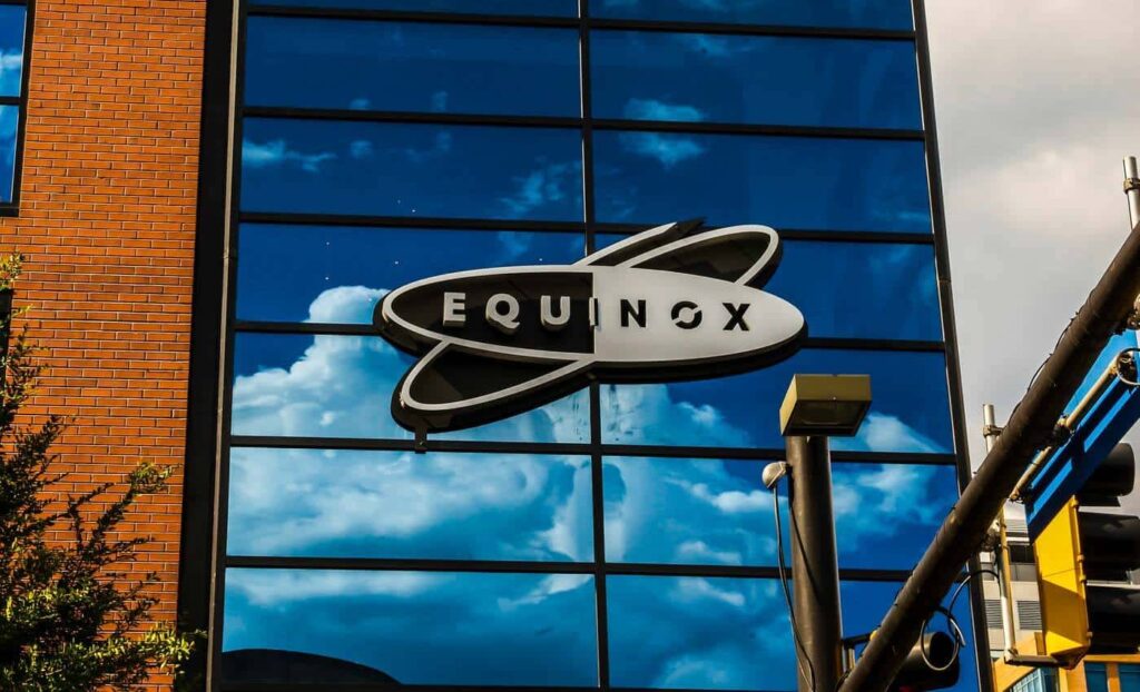 Equinox logo sign displayed on the glass facade of a modern building reflecting clouds and sky.