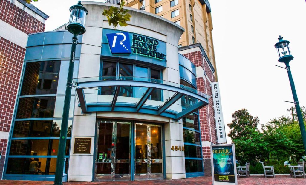 Exterior view of Round House Theatre building with glass entrance, signage, and brick facade at street level.