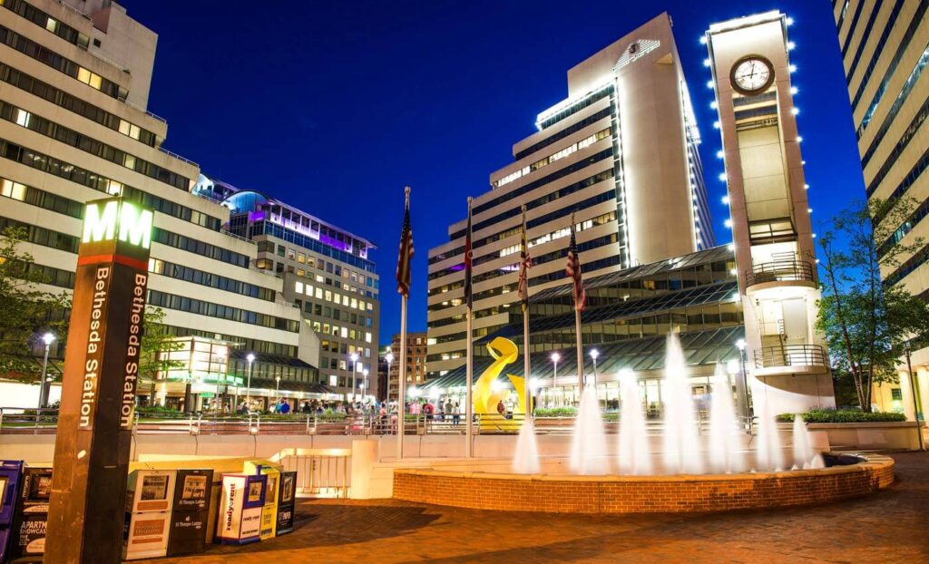 Bethesda Metro Station entrance sign, fountain, modern buildings, clock tower, and lit streetscape at night in Bethesda, Maryland.