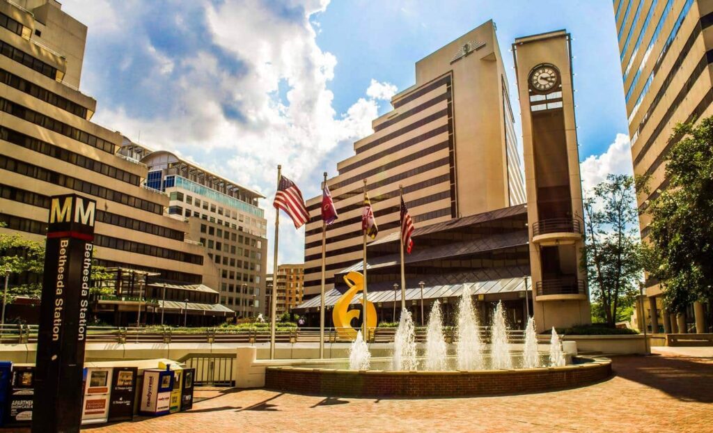 Outdoor plaza with a fountain, flagpoles, a yellow sculpture, and surrounding office buildings; clock tower and Bethesda Metro Station sign visible.