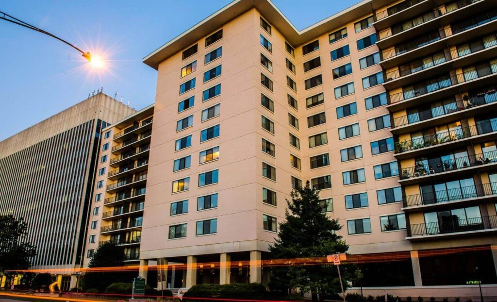 Modern mid-rise apartment building with multiple balconies and windows, photographed at dusk with a streetlight and light trails from passing vehicles in front.