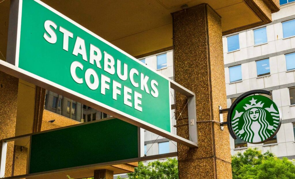 Green Starbucks Coffee sign and round Starbucks logo mounted on the exterior of a building, with windows and greenery in the background.