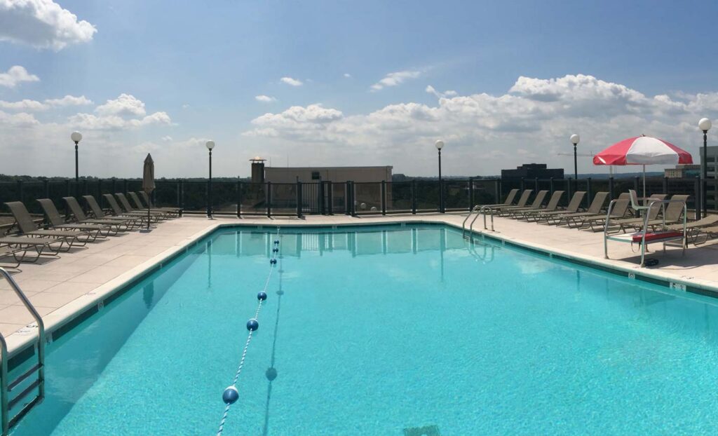 Rooftop swimming pool with clear water, surrounded by lounge chairs and a single red-and-white umbrella under a partly cloudy sky.