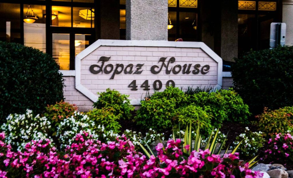 A sign reading "Topaz House 4400" is displayed in front of a building, surrounded by green bushes and pink, white, and purple flowers.