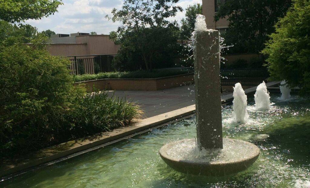 A rectangular stone fountain with multiple water jets is set in a shallow pool, surrounded by greenery and a brick patio area.
