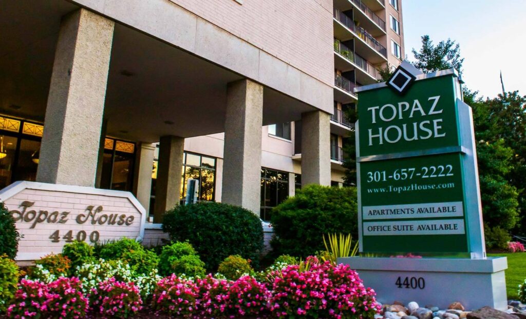 The image shows the entrance of Topaz House apartments at 4400, with a sign listing phone number and availability for apartments and office suites, surrounded by greenery and flowers.